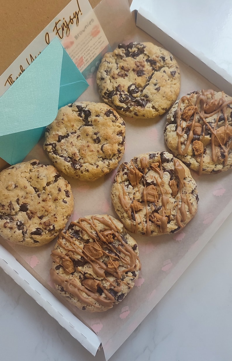 May include: A box of six assorted cookies. The cookies are a mix of chocolate chip and chocolate chip with caramel and chocolate drizzle. The box is lined with white paper with a pink cupcake pattern. The box has a handwritten note that says "Thank You & Enjoy!"