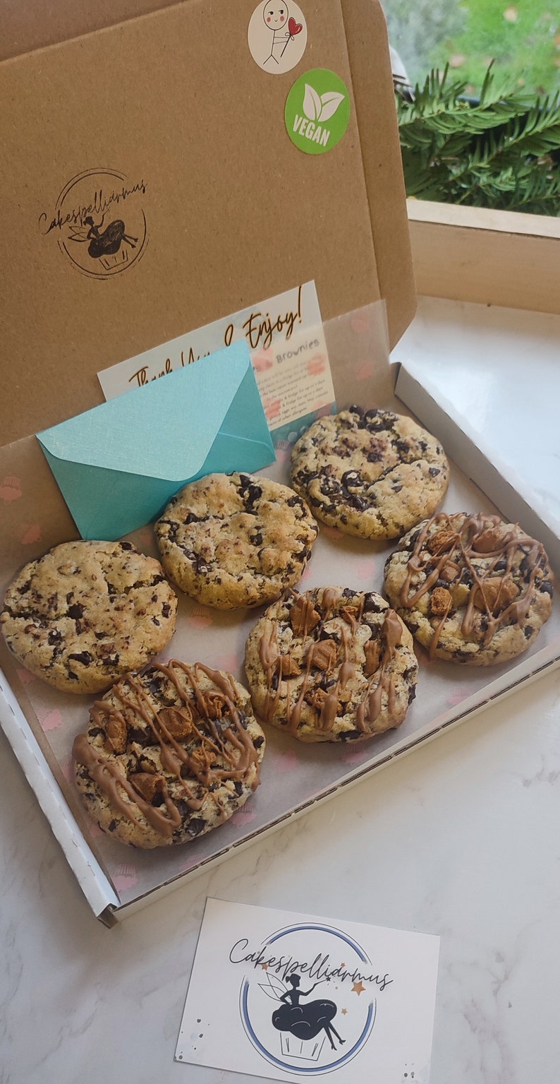 May include: A box of six vegan chocolate chip cookies. The cookies are arranged in a single layer in the box. The box is brown and has a sticker that says "Vegan".