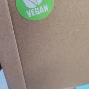 May include: A brown cardboard box with a green sticker that says "VEGAN" with a white leaf design. The box has a clear plastic window showing a cookie.