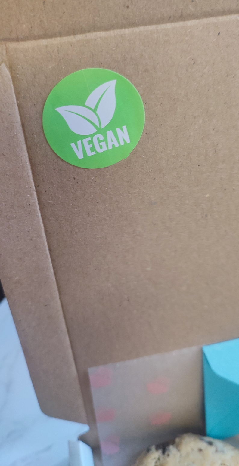 May include: A brown cardboard box with a green sticker that says "VEGAN" with a white leaf design. The box has a clear plastic window showing a cookie.