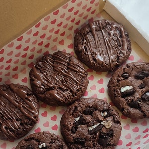 May include: A box of six vegan chocolate chip cookies. The cookies are arranged in a circle on pink and white heart-shaped paper. The box is labeled "VEGAN".