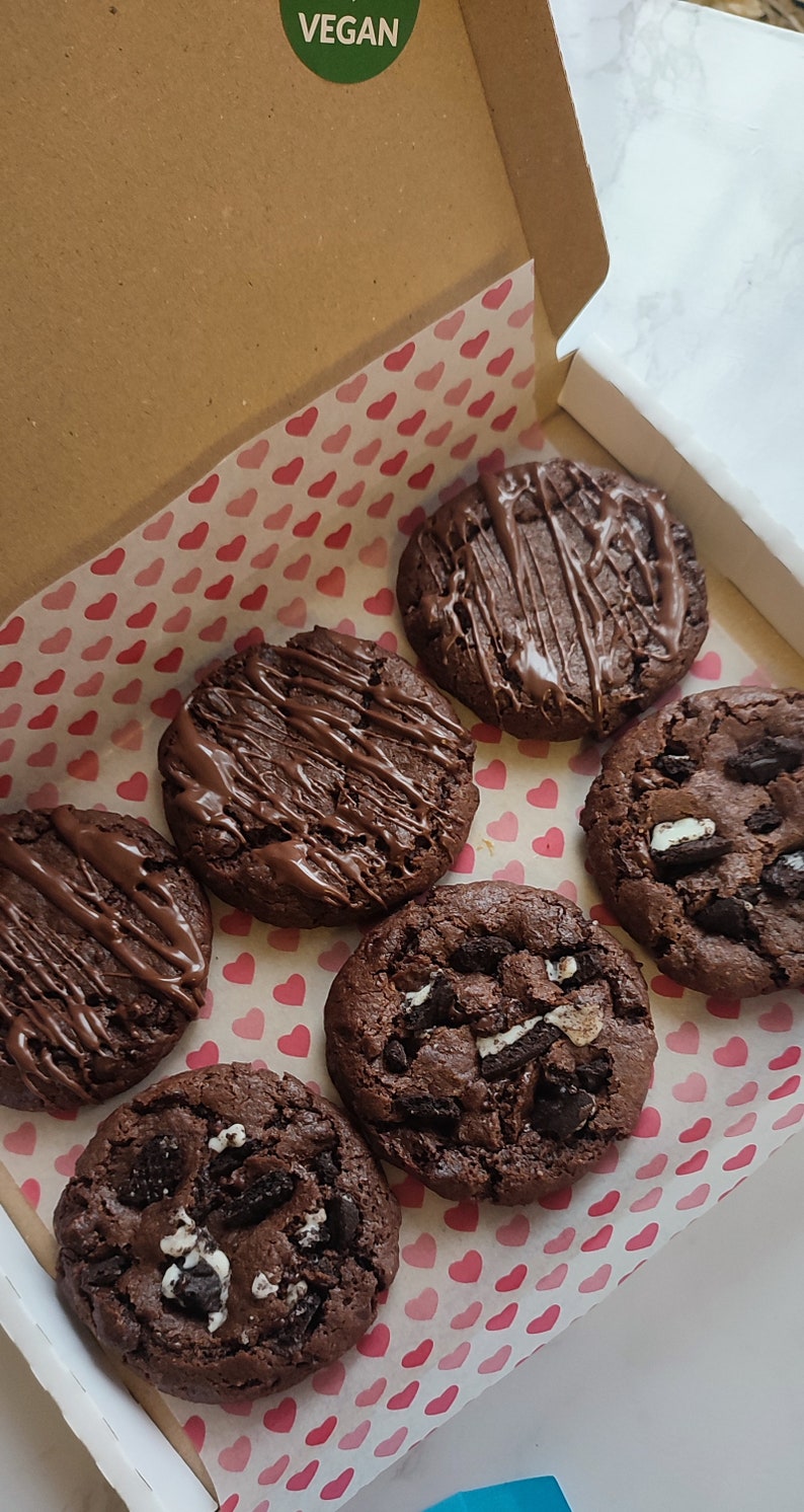 May include: A box of six vegan chocolate chip cookies. The cookies are arranged in a circle on pink and white heart-shaped paper. The box is labeled "VEGAN".