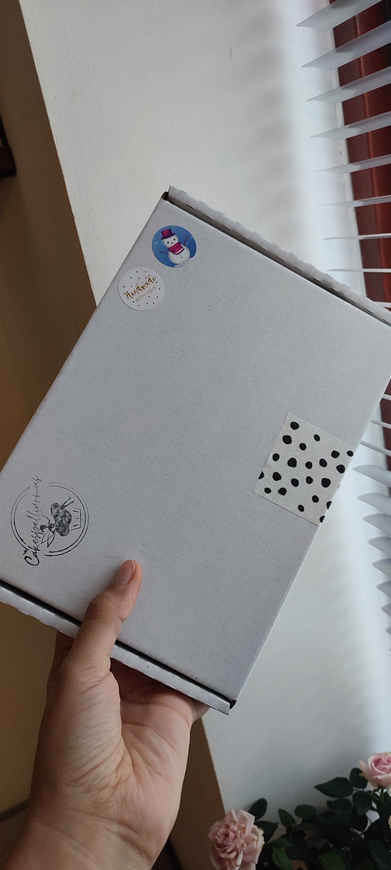 May include: A white cardboard box with a black and white polka dot sticker, a snowman sticker, and a sticker that says "Handmade with Love". The box also has a black and white logo that says "Cakespellings" with a cupcake graphic.