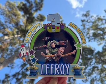 Könnte beinhalten: Ein runder Tortenaufleger mit Buzz Lightyear und Woody aus Toy Story, dem Text To Infinity and Beyond, der Zahl 3 und dem Namen LEEROY. Das Design enthält grüne, blaue und violette Akzente, eine Rakete und zwei grüne Außerirdische.