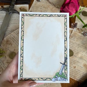 Op de afbeelding: Een rechthoekige briefkaart met een decoratieve rand van groene wijnranken en bloemen. Een illustratie van een zwaard in steen staat in de rechter benedenhoek. De kaart wordt in een hand gehouden, met een zwaard en andere items op de achtergrond.
