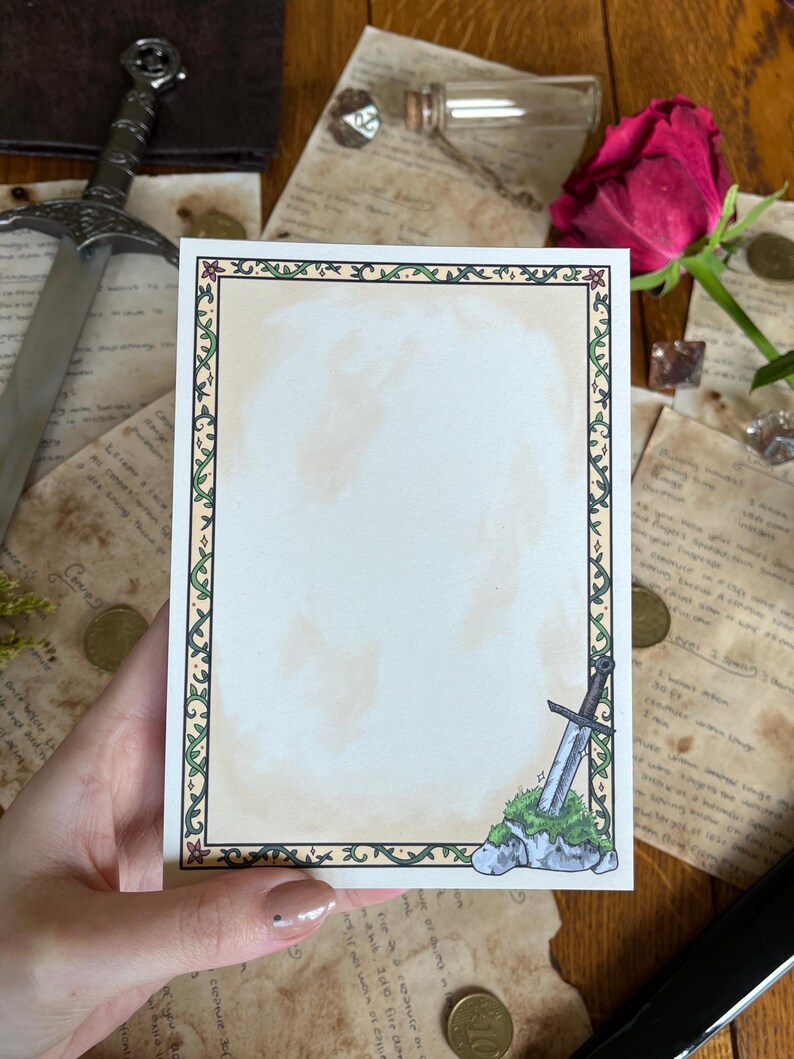 Op de afbeelding: Een rechthoekige briefkaart met een decoratieve rand van groene wijnranken en bloemen. Een illustratie van een zwaard in steen staat in de rechter benedenhoek. De kaart wordt in een hand gehouden, met een zwaard en andere items op de achtergrond.