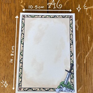 Op de afbeelding: A6-papier, gerecycled en ongestreken, met een decoratieve rand. Het papier meet 10,5 cm x 14,8 cm en bevat 50 pagina's. Een zwaard is in de hoek afgebeeld. Het papier is 100 g/m&sup2;.