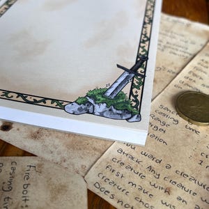 Op de afbeelding: Een notitieblok met een fantasiethema. De hoek van het notitieblok heeft een tekening van een zwaard dat in een met mos bedekte steen is ingebed. Het notitieblok heeft een decoratieve rand met een groen rankenontwerp. De achtergrond bevat handgeschreven notities en een munt.