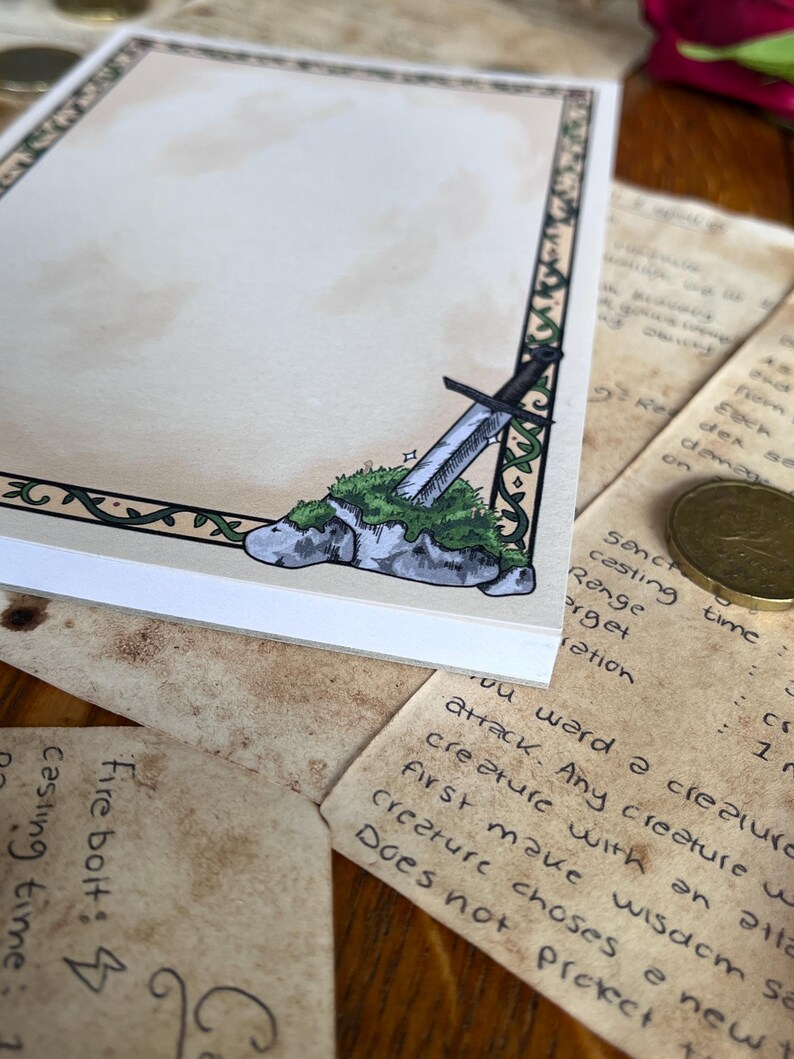 Op de afbeelding: Een notitieblok met een fantasiethema. De hoek van het notitieblok heeft een tekening van een zwaard dat in een met mos bedekte steen is ingebed. Het notitieblok heeft een decoratieve rand met een groen rankenontwerp. De achtergrond bevat handgeschreven notities en een munt.