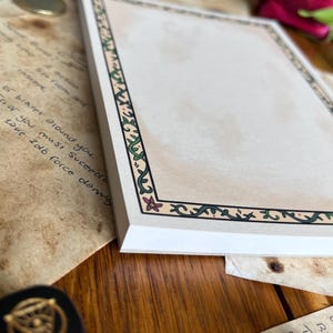 Op de afbeelding: Een stapel blanco papier met een decoratieve rand met groene wijnranken en roze bloemen. Het papier heeft een beige achtergrond en wordt omringd door andere items, waaronder handgeschreven notities en een zwarte dobbelsteen.