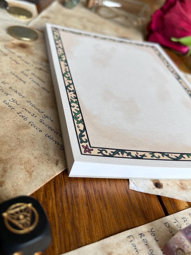 Op de afbeelding: Een stapel blanco papier met een decoratieve rand met groene wijnranken en roze bloemen. Het papier heeft een beige achtergrond en wordt omringd door andere items, waaronder handgeschreven notities en een zwarte dobbelsteen.