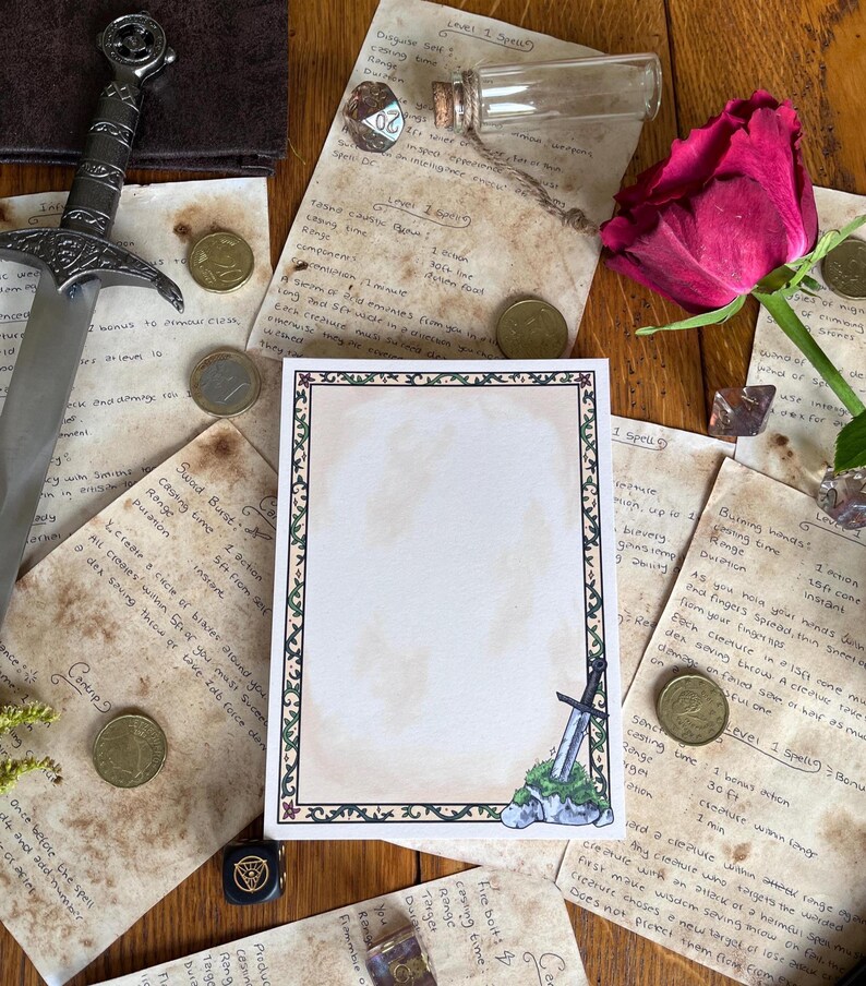 Op de afbeelding: Een artistieke compositie met een fantasiethema. Een decoratief papier met rand is omgeven door handgeschreven notities, munten, een zwaard, een roos en een kleine glazen flacon. Het papier heeft een tekening van een zwaard.