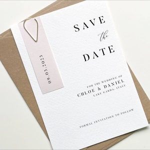 Puede incluir: Tarjeta de guardar la fecha con una etiqueta rosa adjunta. La tarjeta dice "Save the Date" en negro con un "the" escrito a mano. La etiqueta dice "09.03.2023" en negro. La tarjeta tambi&eacute;n dice "For the wedding of Chloe & Daniel, Lake Garda, Italy" y "Formal invitation to follow".