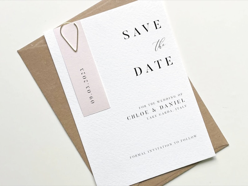 Puede incluir: Tarjeta de guardar la fecha con una etiqueta rosa adjunta. La tarjeta dice "Save the Date" en negro con un "the" escrito a mano. La etiqueta dice "09.03.2023" en negro. La tarjeta tambi&eacute;n dice "For the wedding of Chloe & Daniel, Lake Garda, Italy" y "Formal invitation to follow".