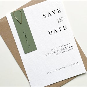 Puede incluir: Tarjeta de guardar la fecha con una etiqueta verde adjunta. La tarjeta dice "Save the Date" en texto negro con un "the" escrito a mano. El texto debajo dice "For the wedding of Chloe & Daniel, Lake Garda, Italy" y "Formal invitation to follow". La etiqueta dice "09.03.2023" en texto negro.