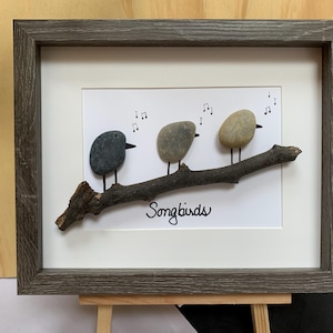 May include: Framed artwork featuring three pebble birds perched on a branch. The birds are various shades of gray and tan, with black beaks and legs. The branch is brown, and the background is white. The word "Songbirds" is written below the branch.