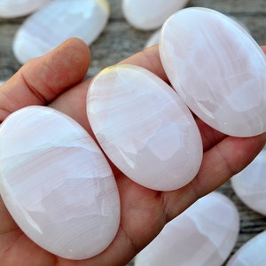 Puede incluir: Una mano sostiene varias piedras rosas lisas de forma ovalada con un brillo nacarado. Las piedras tienen un aspecto suave y transl&uacute;cido con sutiles vetas blancas. Se ven m&aacute;s piedras en el fondo.