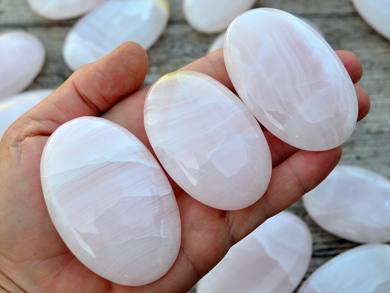 Puede incluir: Una mano sostiene varias piedras rosas lisas de forma ovalada con un brillo nacarado. Las piedras tienen un aspecto suave y transl&uacute;cido con sutiles vetas blancas. Se ven m&aacute;s piedras en el fondo.