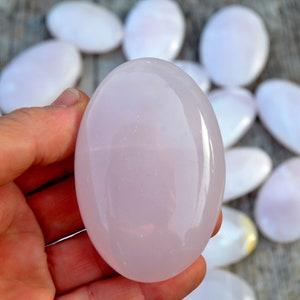 Puede incluir: Una mano sostiene una piedra rosa grande y lisa de forma ovalada, con varias piedras similares dispersas en el fondo. Las piedras tienen un aspecto pulido y brillante y var&iacute;an ligeramente en tama&ntilde;o y color, con un suave tono rosa p&aacute;lido.