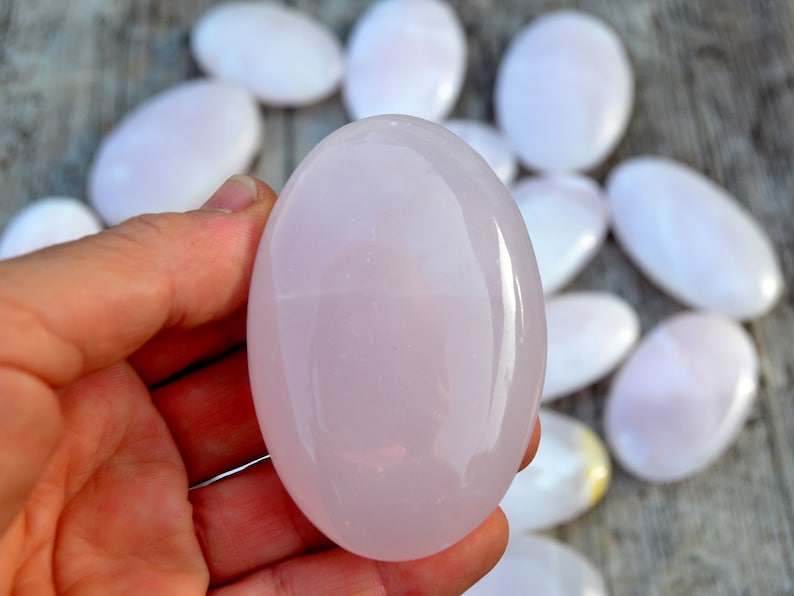Puede incluir: Una mano sostiene una piedra rosa grande y lisa de forma ovalada, con varias piedras similares dispersas en el fondo. Las piedras tienen un aspecto pulido y brillante y var&iacute;an ligeramente en tama&ntilde;o y color, con un suave tono rosa p&aacute;lido.