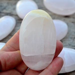 Puede incluir: Un conjunto de piedras lisas de forma ovalada en tonos blancos y rosa p&aacute;lido. Una piedra se sostiene en la mano, mostrando su superficie pulida. Las piedras tienen un aspecto natural y org&aacute;nico, con sutiles variaciones de color y textura. Medidas en cent&iacute;metros.