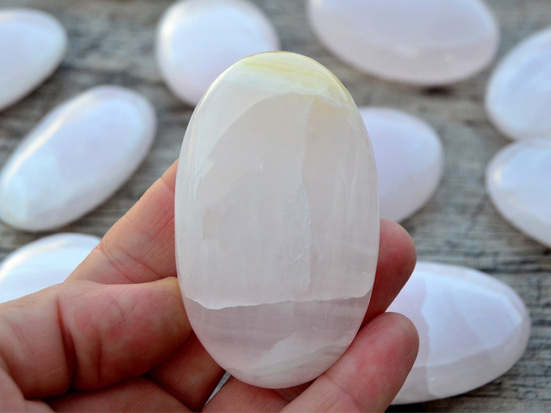 Puede incluir: Un conjunto de piedras lisas de forma ovalada en tonos blancos y rosa p&aacute;lido. Una piedra se sostiene en la mano, mostrando su superficie pulida. Las piedras tienen un aspecto natural y org&aacute;nico, con sutiles variaciones de color y textura. Medidas en cent&iacute;metros.