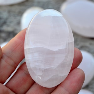 Puede incluir: Una piedra pulida, de forma ovalada, de color rosa p&aacute;lido, sostenida en una mano. La piedra tiene una superficie lisa y brillante con patrones sutiles y arremolinados. Otras piedras similares son visibles en el fondo.
