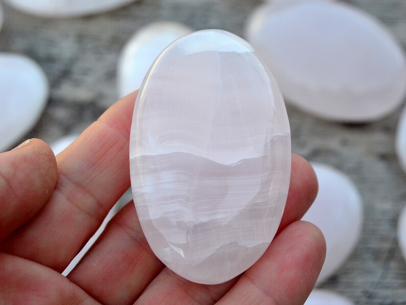 Puede incluir: Una piedra pulida, de forma ovalada, de color rosa p&aacute;lido, sostenida en una mano. La piedra tiene una superficie lisa y brillante con patrones sutiles y arremolinados. Otras piedras similares son visibles en el fondo.