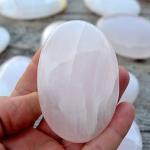 Puede incluir: Una mano sostiene una piedra lisa, ovalada, de color rosa p&aacute;lido. Otras piedras similares est&aacute;n dispersas en el fondo. Las piedras tienen un aspecto pulido y brillante, y el color es un rosa suave y apagado.