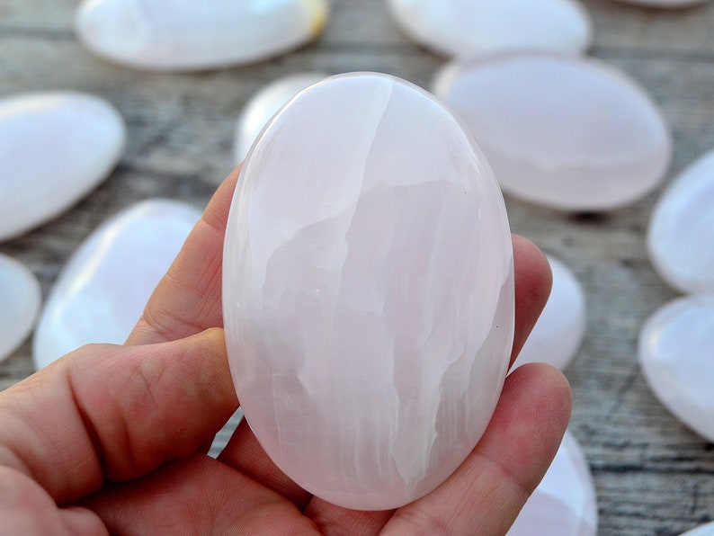 Puede incluir: Una mano sostiene una piedra lisa, ovalada, de color rosa p&aacute;lido. Otras piedras similares est&aacute;n dispersas en el fondo. Las piedras tienen un aspecto pulido y brillante, y el color es un rosa suave y apagado.