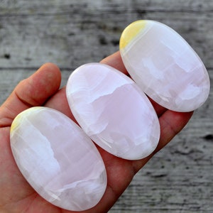 Puede incluir: Tres piedras ovaladas pulidas en tonos rosa y blanco, con un toque de amarillo en la parte superior. Las piedras son lisas y se sostienen en una mano, mostrando su belleza natural. El fondo es una superficie de madera.