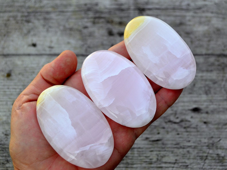 Puede incluir: Tres piedras ovaladas pulidas en tonos rosa y blanco, con un toque de amarillo en la parte superior. Las piedras son lisas y se sostienen en una mano, mostrando su belleza natural. El fondo es una superficie de madera.