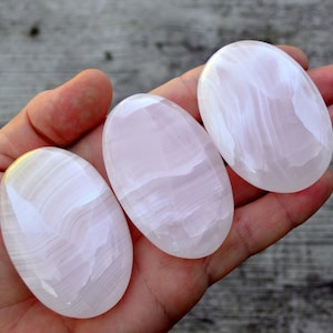 Puede incluir: Tres piedras ovaladas pulidas, en tonos rosas y blancos, se sostienen en una mano. Las piedras tienen una superficie lisa y brillante con sutiles vetas. Las piedras miden aproximadamente 7.6 cm de largo.
