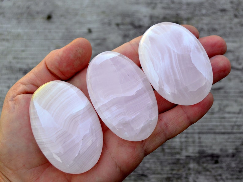 Puede incluir: Tres piedras ovaladas pulidas, en tonos rosas y blancos, se sostienen en una mano. Las piedras tienen una superficie lisa y brillante con sutiles vetas. Las piedras miden aproximadamente 7.6 cm de largo.
