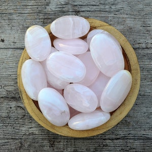 Puede incluir: Un cuenco de madera lleno de piedras rosas lisas y ovaladas. Las piedras tienen un aspecto pulido y diferentes tonos de rosa. El cuenco est&aacute; sobre una superficie de madera envejecida, creando una est&eacute;tica natural.