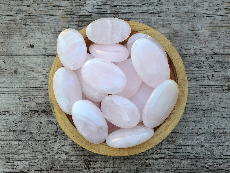 Puede incluir: Un cuenco de madera lleno de piedras rosas lisas y ovaladas. Las piedras tienen un aspecto pulido y diferentes tonos de rosa. El cuenco est&aacute; sobre una superficie de madera envejecida, creando una est&eacute;tica natural.