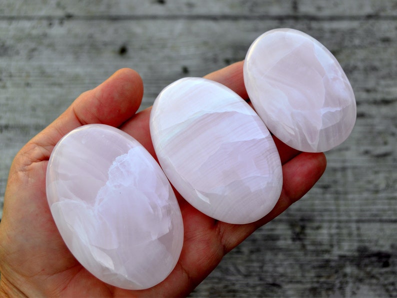 Puede incluir: Tres piedras ovaladas pulidas en tonos rosa y blanco. Las piedras tienen una superficie lisa y brillante con patrones de vetas naturales. Las piedras se sostienen en una mano, mostrando sus variaciones de tama&ntilde;o y color. Las piedras probablemente se utilizan con fines decorativos o espirituales.