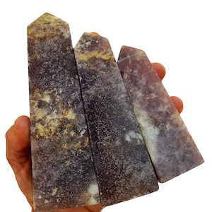 Op de afbeelding: Drie paarse en bruine kristallen obelisken met een textuur worden in een hand gehouden. De obelisken zijn rechthoekig met een puntige top. De kristallen hebben een mix van paarse, bruine en witte kleuren.