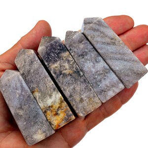 Op de afbeelding: Zes rechthoekige, grijs-paarse kristallen met een gepolijste, gestreepte uitstraling worden in een hand gehouden. De kristallen hebben een natuurlijk, getextureerd oppervlak met kleurvariaties, waaronder hints van geel en wit.
