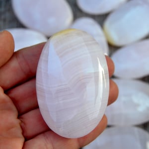 Puede incluir: Una piedra pulida, de forma ovalada, de color rosa p&aacute;lido, con sutiles vetas blancas. La piedra se sostiene en una mano, con varias piedras similares visibles en el fondo. La superficie es lisa y reflectante.