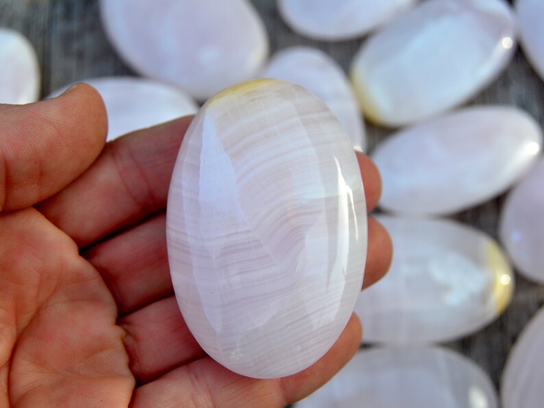 Puede incluir: Una piedra pulida, de forma ovalada, de color rosa p&aacute;lido, con sutiles vetas blancas. La piedra se sostiene en una mano, con varias piedras similares visibles en el fondo. La superficie es lisa y reflectante.