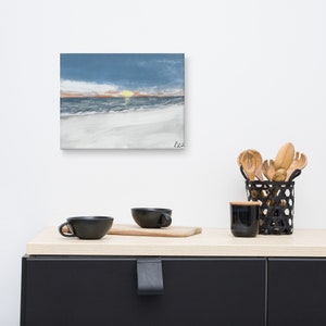 May include: A painting depicting a beach scene with a sunset over the ocean. The sky is blue and orange, and the water is a light blue. The sand is white. The painting is on a white wall, and there are some kitchen items on a counter in front of it.