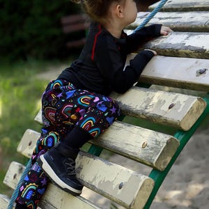 May include: A child in black leggings with a colourful rainbow and heart pattern, black shoes, and a black long-sleeved top with red accents. The child is climbing on a wooden structure.