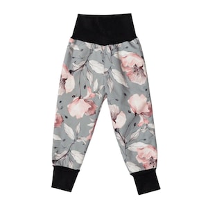 Op de afbeelding: Grijze broek met bloemenprint, hoge zwarte tailleband en zwarte manchetten. De broek heeft een patroon van roze bloemen en witte bladeren. De stof lijkt zacht en comfortabel, geschikt voor vrijetijdskleding.