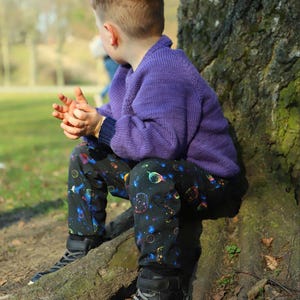 Puede incluir: Un niño pequeño con un suéter morado y pantalones negros con estampado espacial se sienta en un tronco de árbol. Lleva zapatillas negras de caña alta.