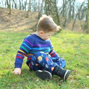 May include: A young child wearing a blue and rainbow striped sweater and blue pants with a planet pattern sits on the grass in a forest.