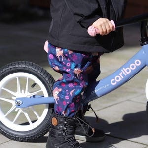 Può includere: Un bambino con pantaloni neri con stampa di cervo e fiori cavalca una bicicletta a bilanciamento blu e bianca con il marchio "cariboo" e "LEDventure" stampato sul telaio. Il bambino indossa stivali invernali neri.