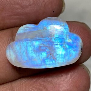 May include: A cloud-shaped moonstone gemstone with a blue and white iridescent glow. The polished stone is held in a hand, showcasing its natural beauty and unique form. This gemstone is ideal for jewelry making or collecting.