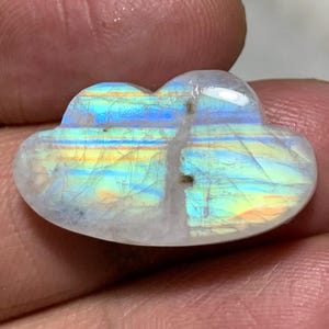 Könnte beinhalten: Ein wolkenförmiger Edelstein mit Regenbogen-Irisieren. Der polierte Stein ist weiß mit Streifen in Blau, Gelb und Grün. Der Edelstein wird in einer Hand gehalten, was seine einzigartige Form und Farbvariationen zeigt.