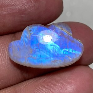 May include: A polished, cloud-shaped moonstone gemstone. The stone has a pearlescent, iridescent sheen with flashes of blue and white. The gemstone is held in a hand, showcasing its unique shape and color.